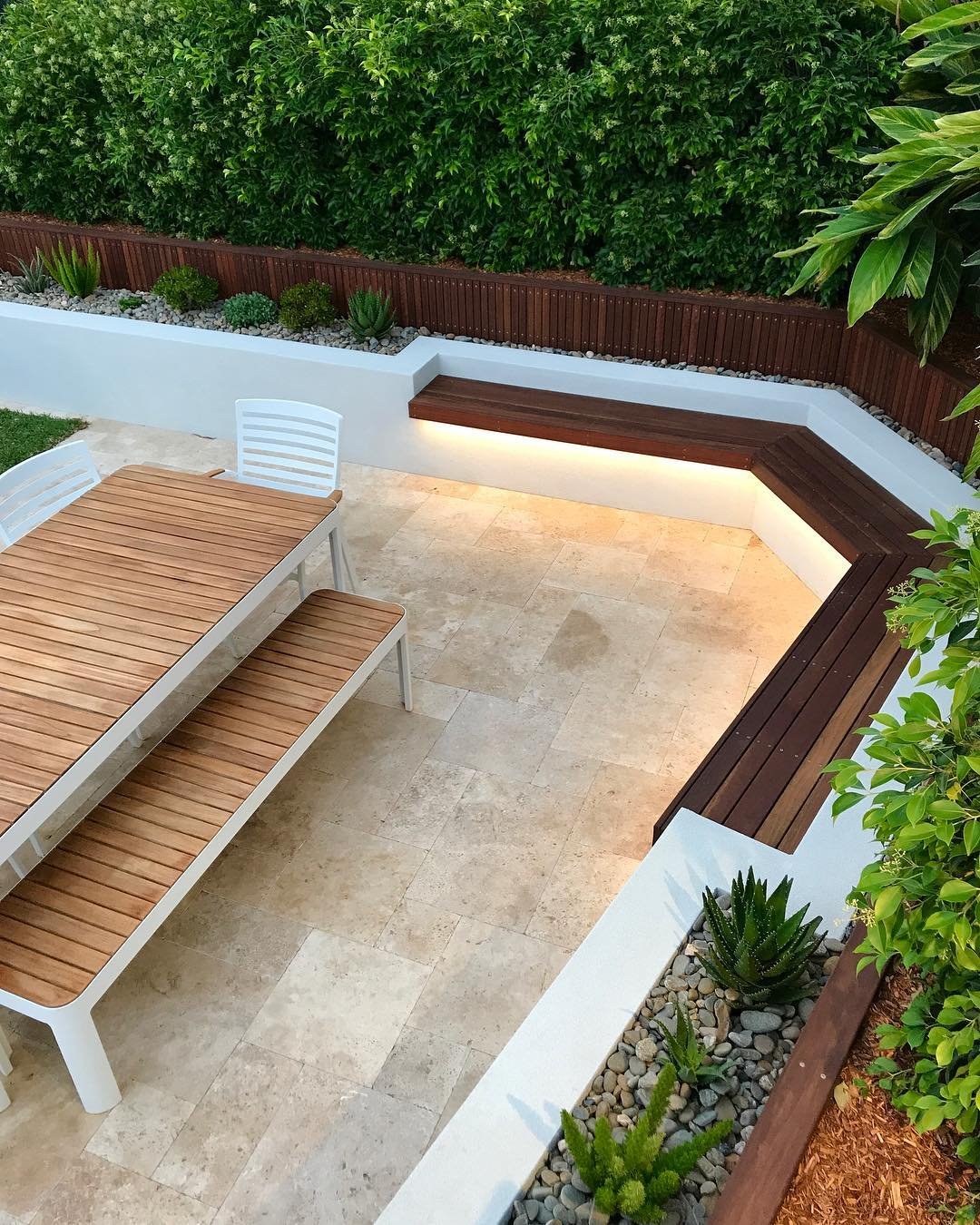Área de jantar externa com piso de travertino em paginação francesa, banco de madeira embutido no muro e iluminação rasante