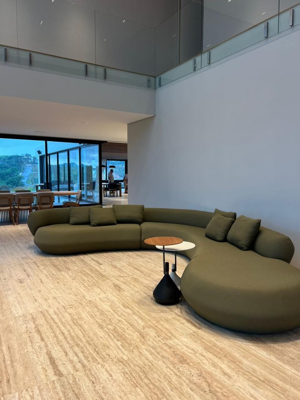 Living de pé-direito duplo com piso contínuo de travertino romano claro, sofá curvo verde-oliva e abertura de vidro para o vale