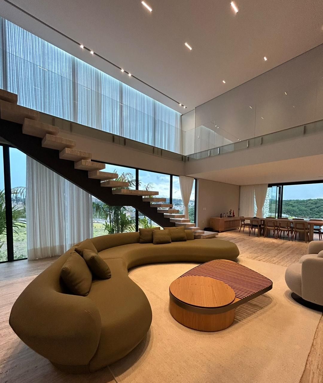 Sala de pé-direito duplo com escada de degraus de travertino em balanço, sofá curvo e vidro do piso ao teto