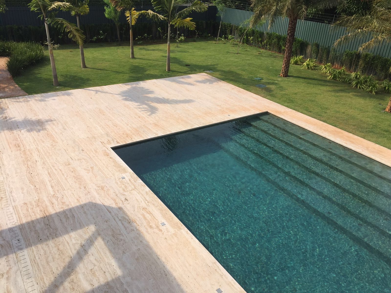 Deck de travertino em placas grandes ao redor de piscina retangular, com sombra de palmeira sobre a pedra