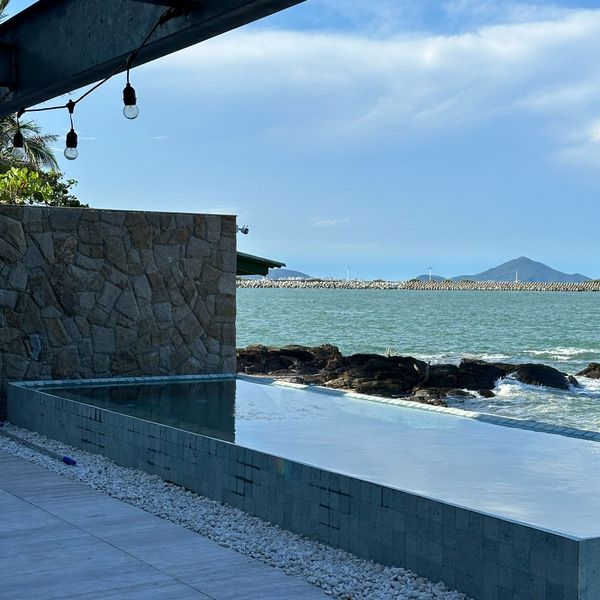 Piscina de borda infinita revestida em Pedra Hijau Lisa, com a face externa em peças verdes e o mar ao fundo