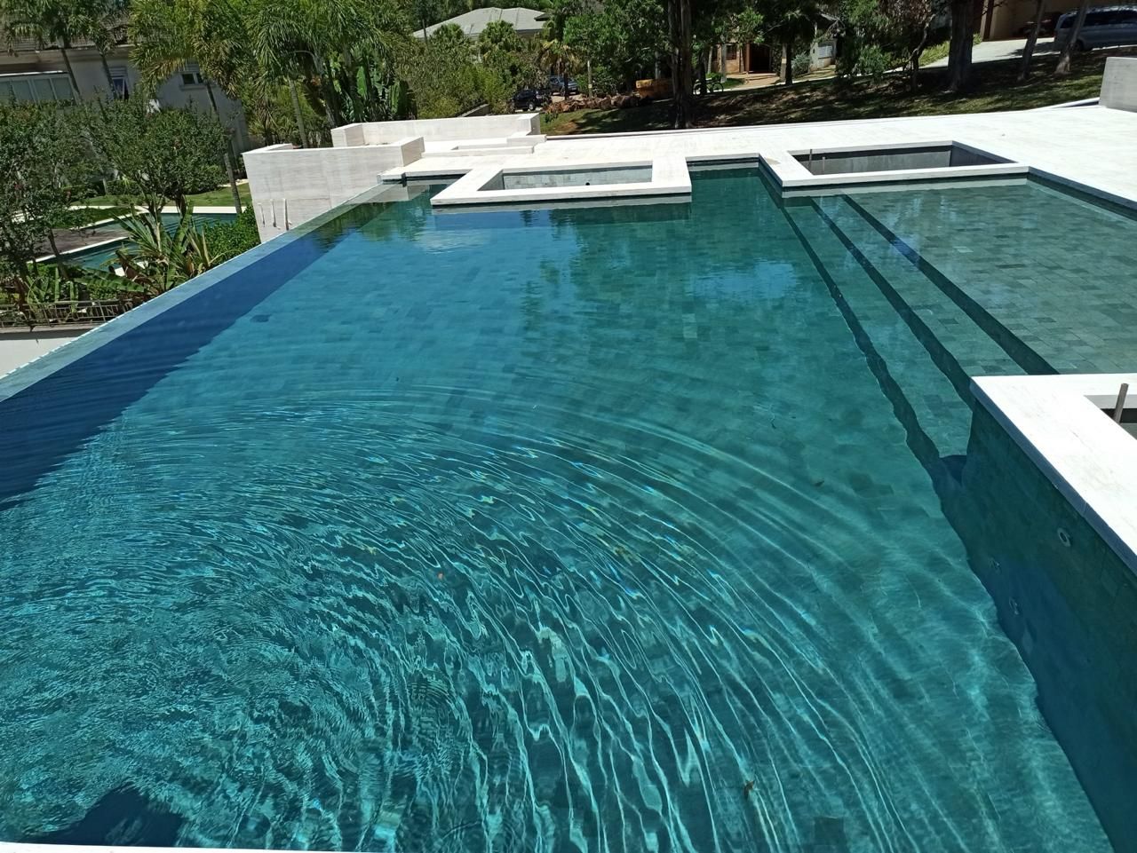 Piscina de borda infinita revestida em pedra verde, com a água em tom turquesa e deck de travertino claro