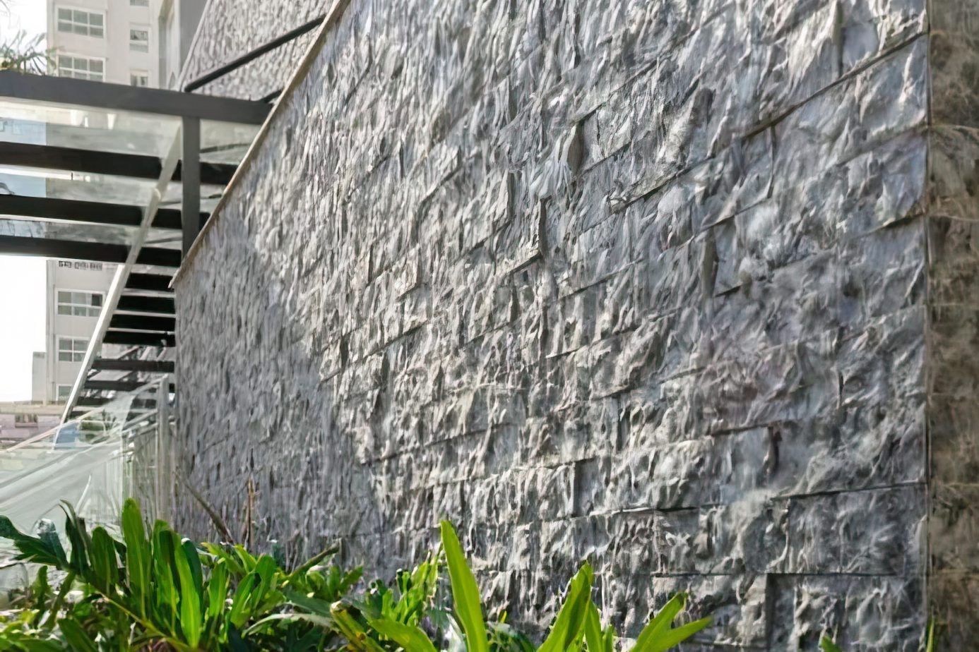 Muro alto revestido em rock face cinza, visto em perspectiva rasante, com jardim na base e prédios ao fundo