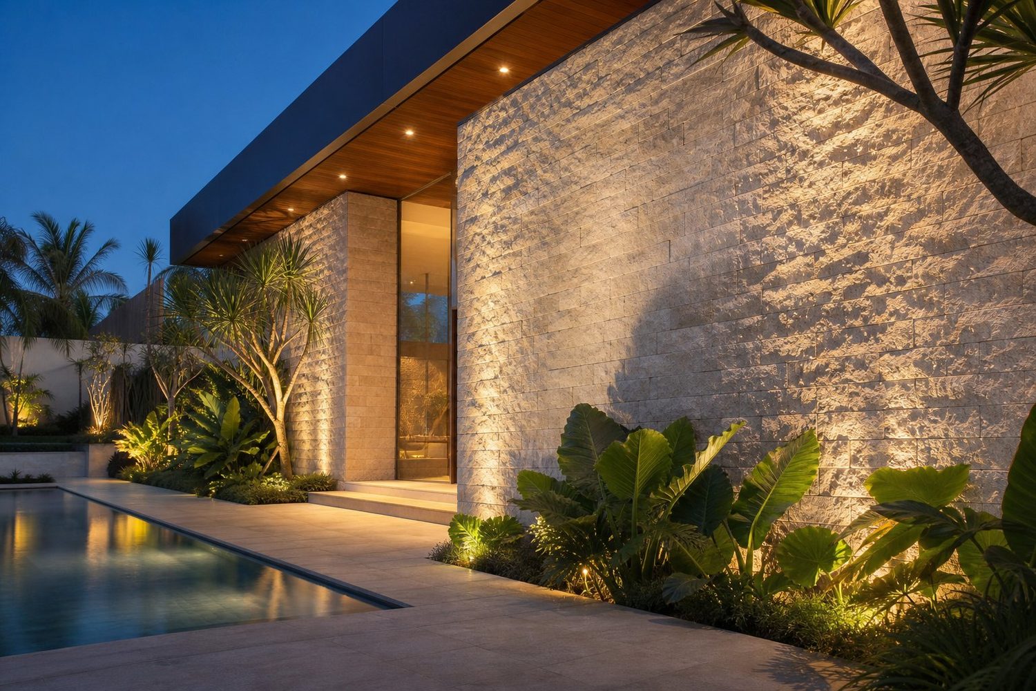Fachada de casa de alto padrão revestida em travertino rock face, iluminada de baixo para cima ao anoitecer, com piscina e jardim tropical