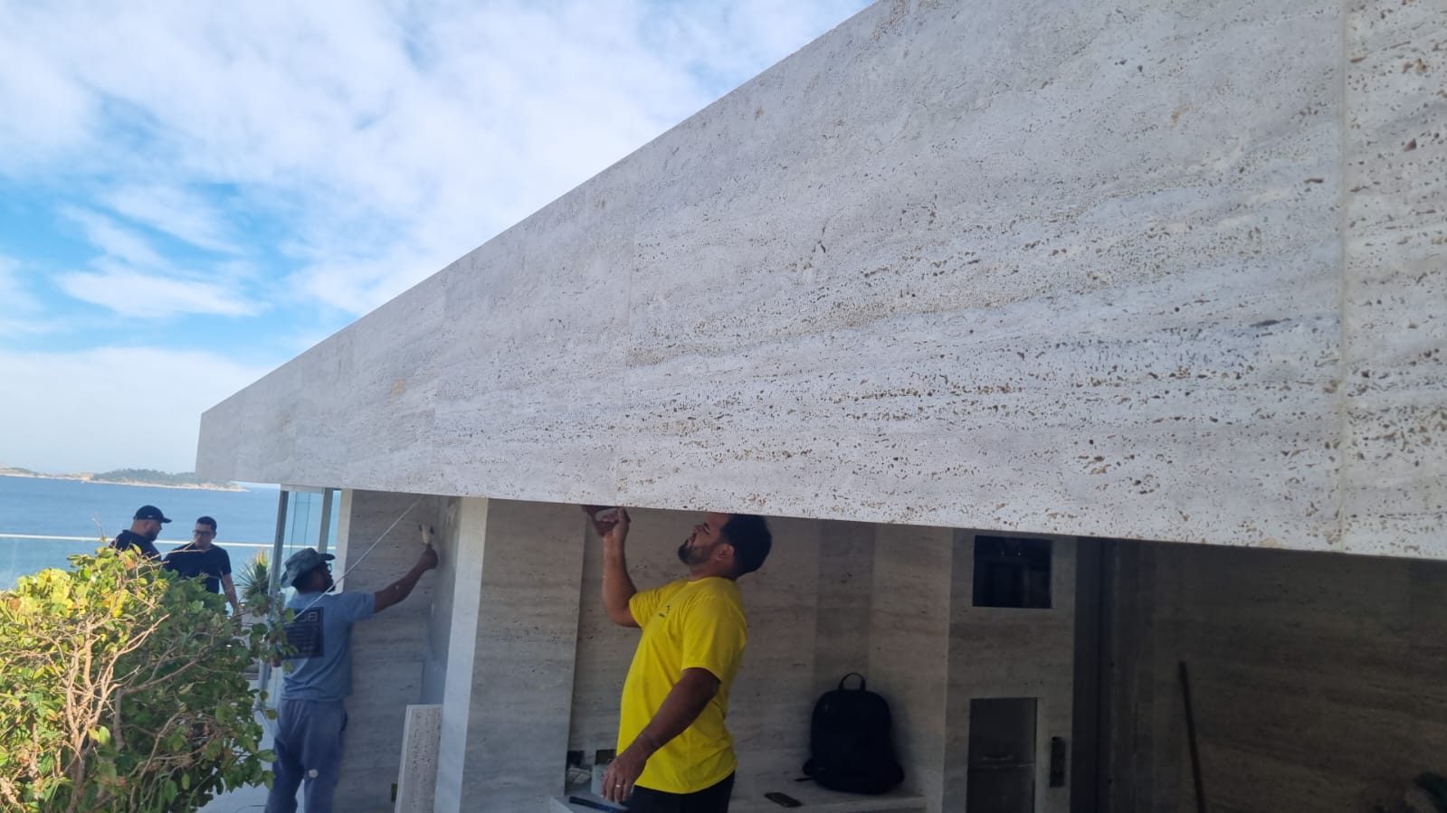 Equipe assentando placas de travertino na fachada de uma casa à beira-mar, com a peça apoiada durante a colagem