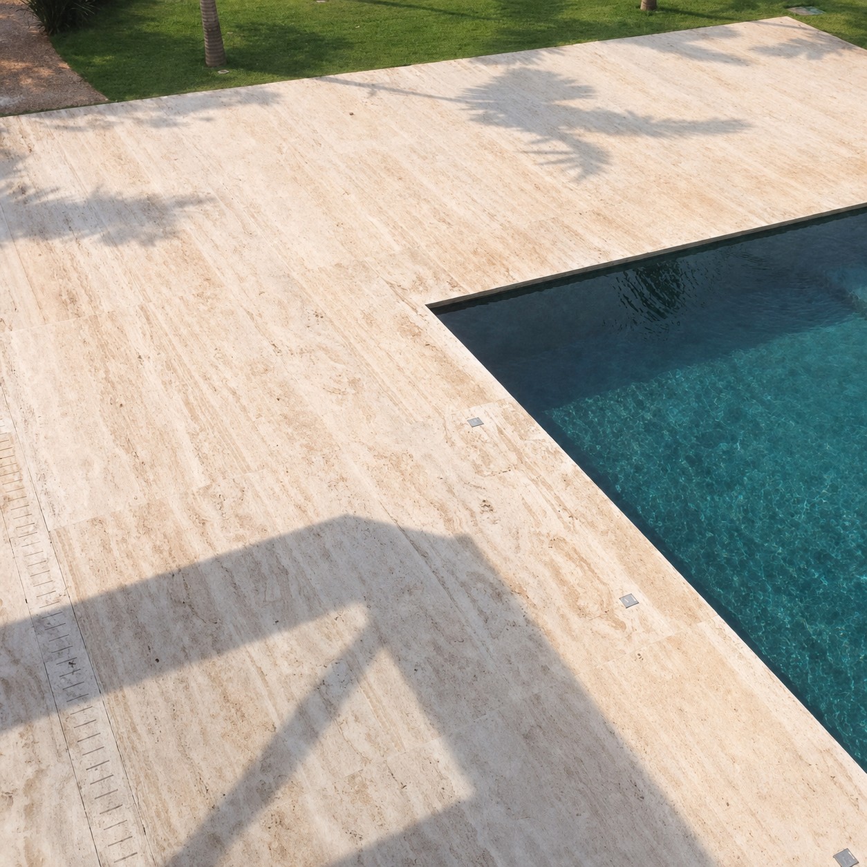 Deck de travertino claro ao redor de uma piscina, visto de cima, com grama e sombra de palmeira sobre a pedra